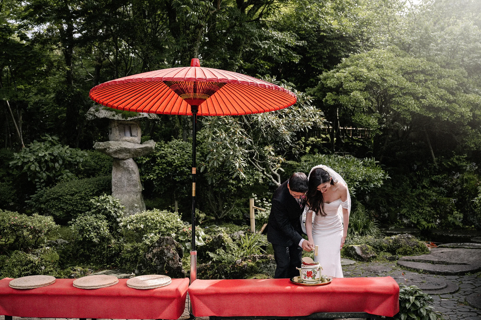 The couple cut their cake beneath a red paper parasol at a garden table draped in red, a stone lantern behind them.