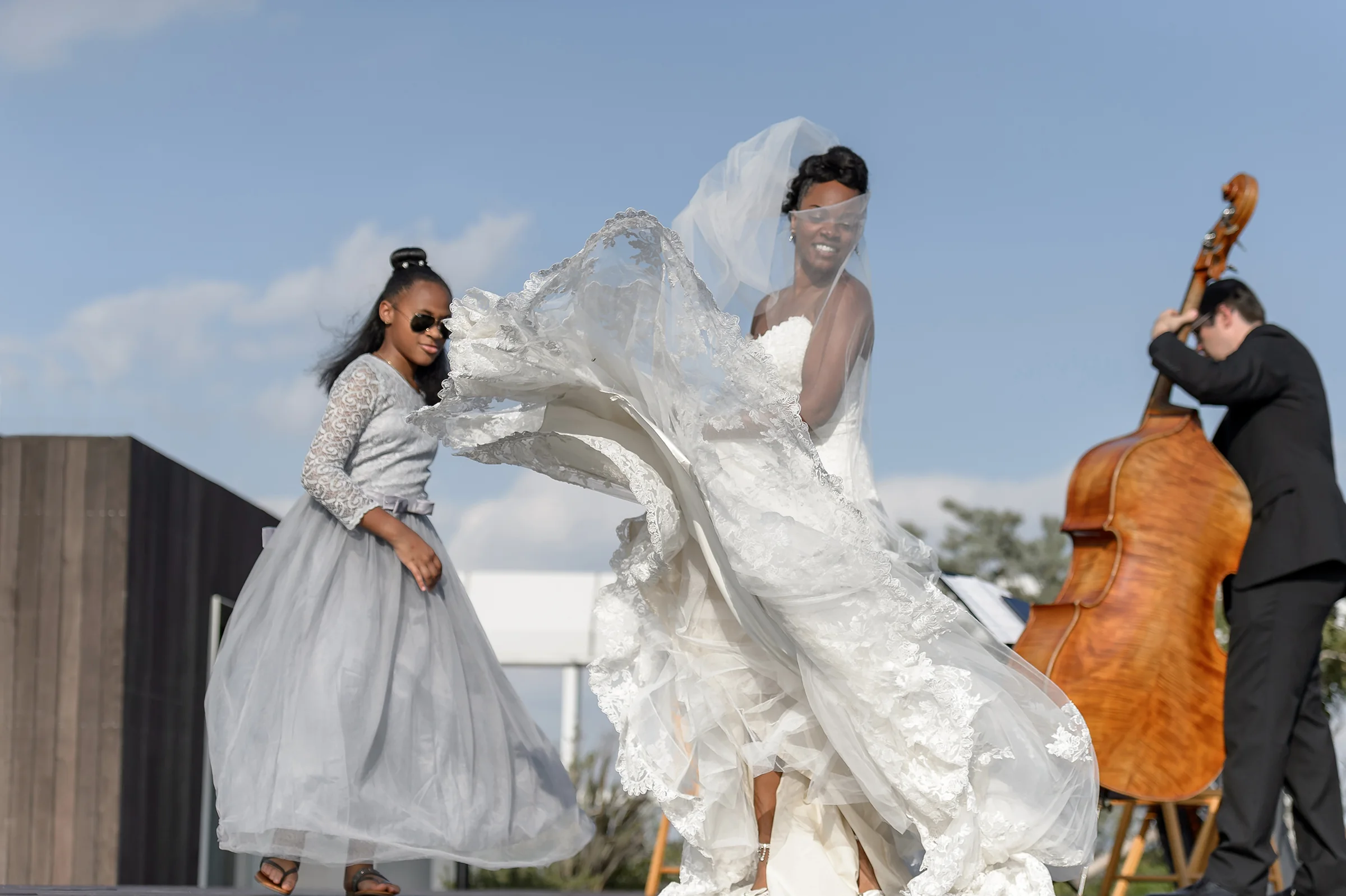 The bride dances in the wind beside a double bass player on a rooftop, her veil flying.
