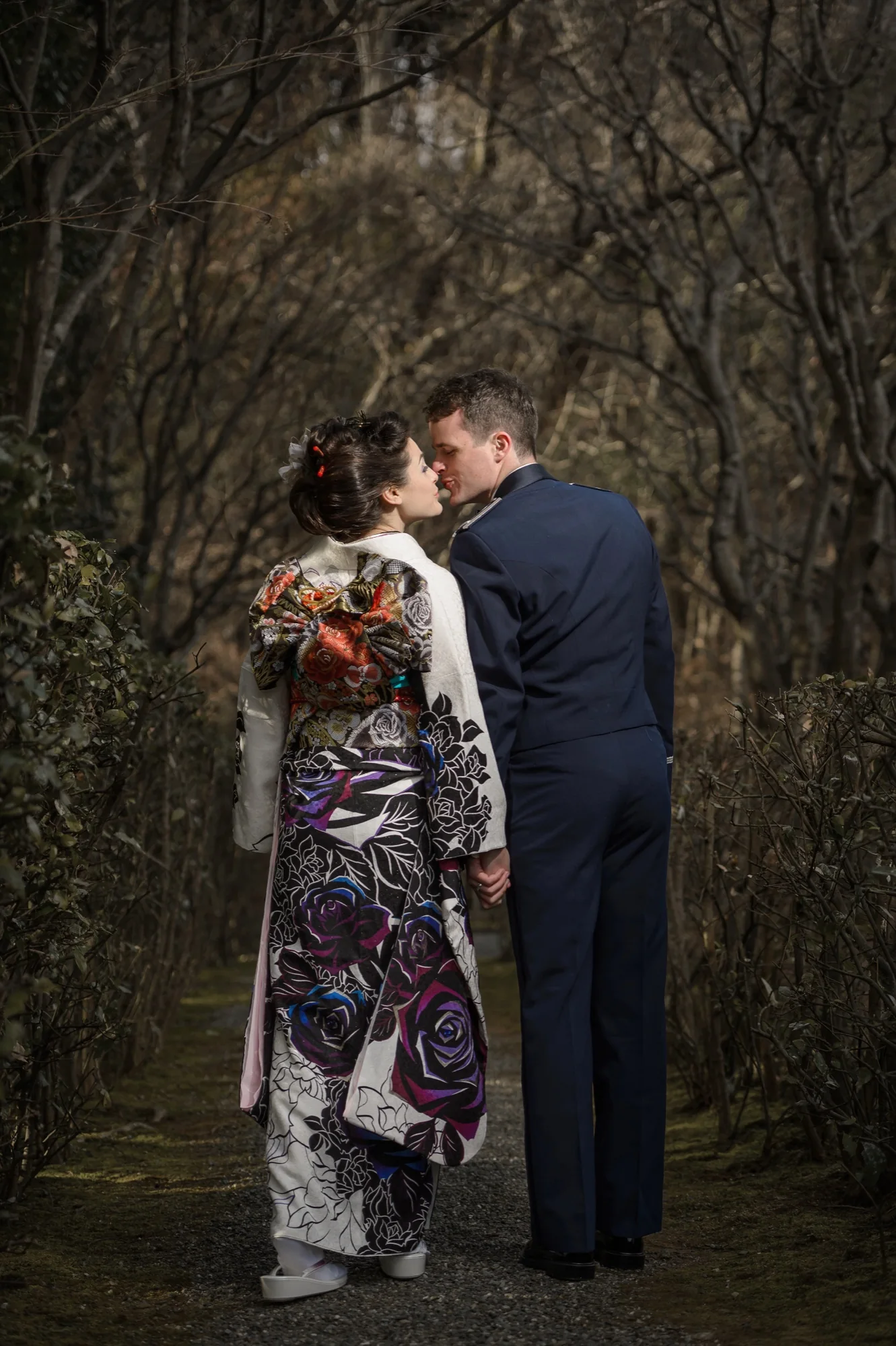 Seen from behind on a garden path, the bride in a rose-patterned furisode and the groom in uniform share a kiss.