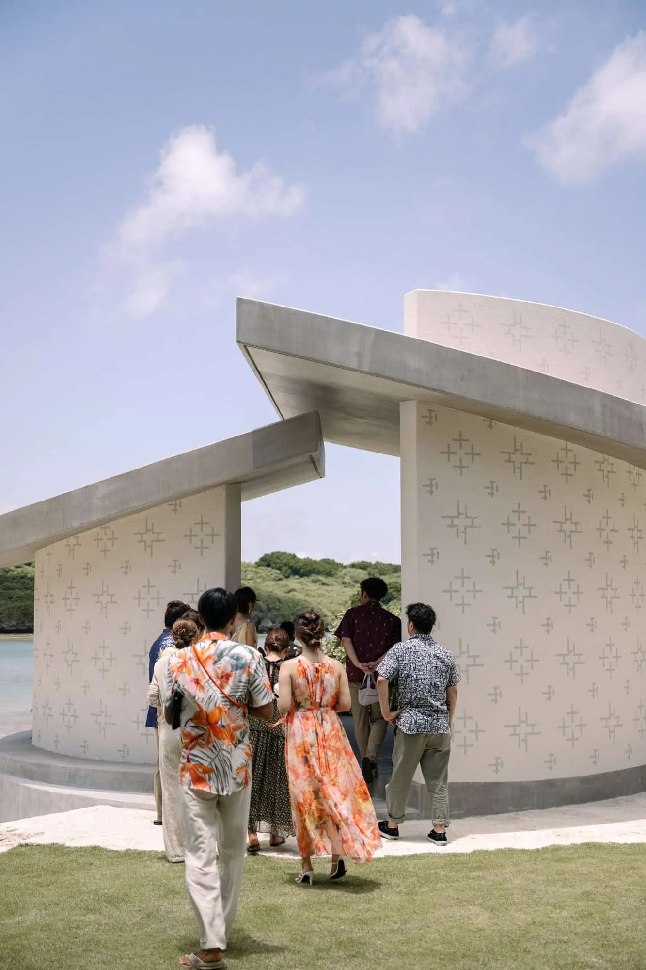 Guests in floral shirts and a long orange dress walk into the round chapel for a Rosewood Miyakojima wedding, a green headland across the water.