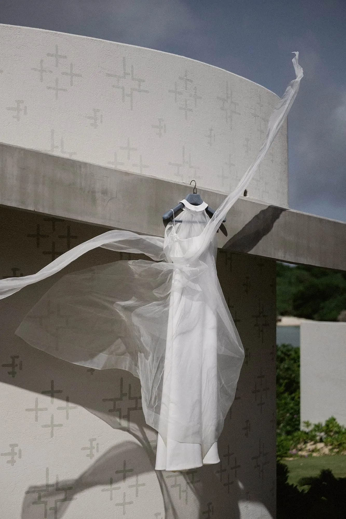 The bride's white halter dress hangs on the curved, patterned chapel wall on the morning of her Rosewood Miyakojima wedding, lifting in the wind.