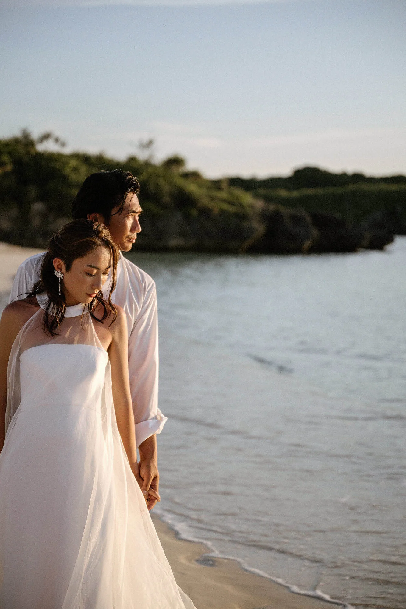 The bride and groom stand close on a Miyakojima beach in golden evening light, a low green headland across the calm water behind them.