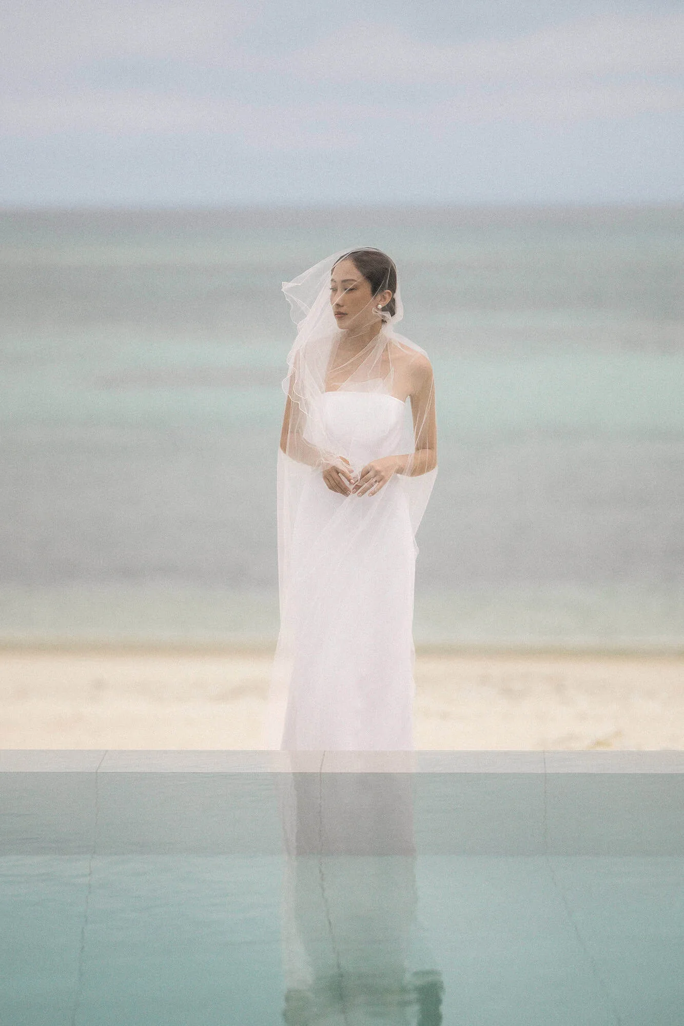 A bridal portrait at the infinity pool, her veil wrapped round her face and her reflection below, the day before her wedding in Okinawa.