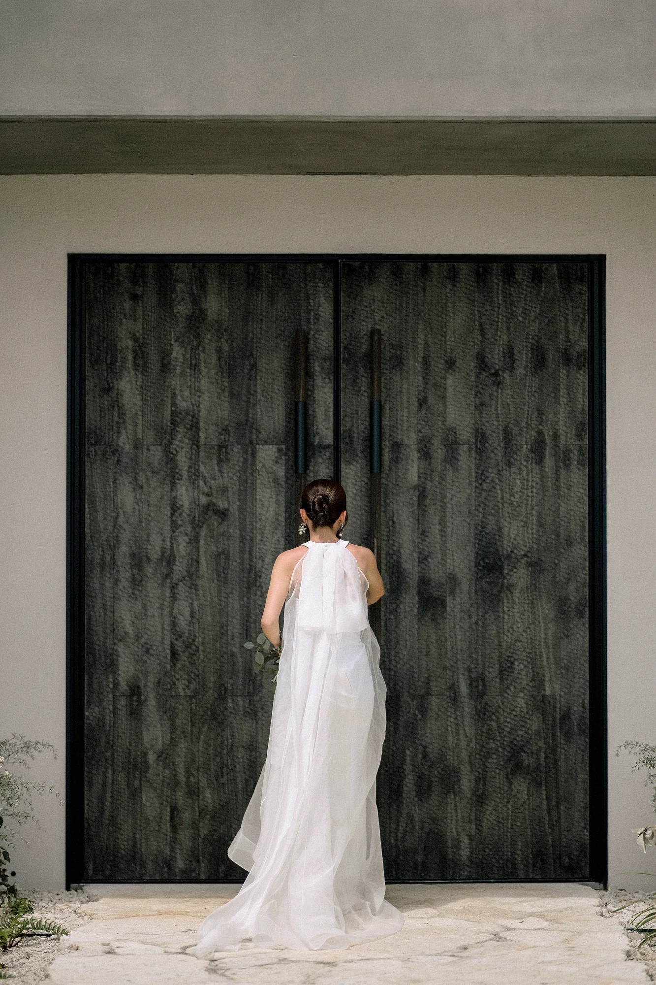 Seen from behind, the bride stands before a pair of tall dark timber doors at Rosewood Miyakojima, her silk cape falling to the stone.