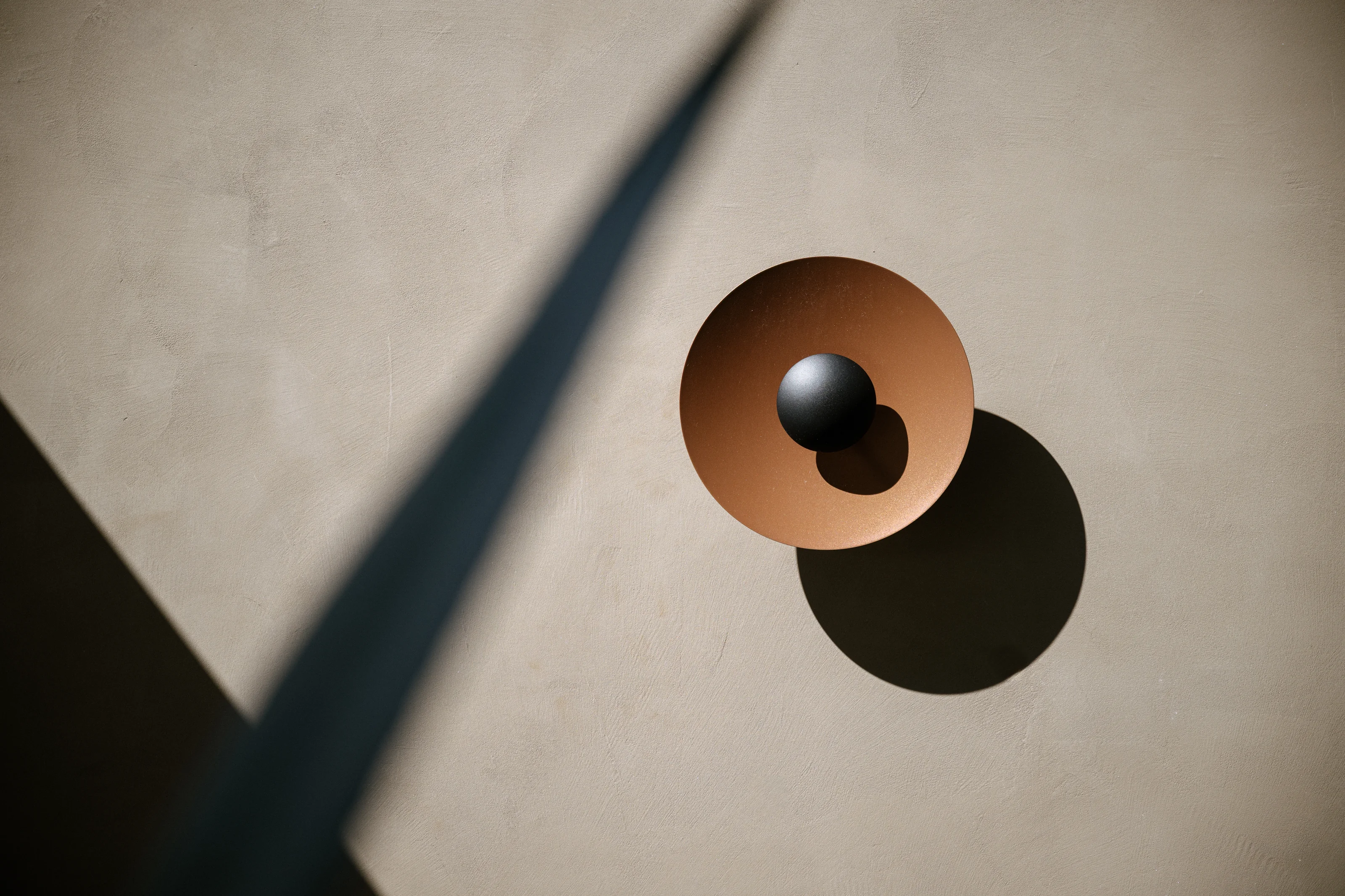 A copper sphere and its shadow on a pale wall, crossed by the dark blade of a leaf, at Rosewood Miyakojima.