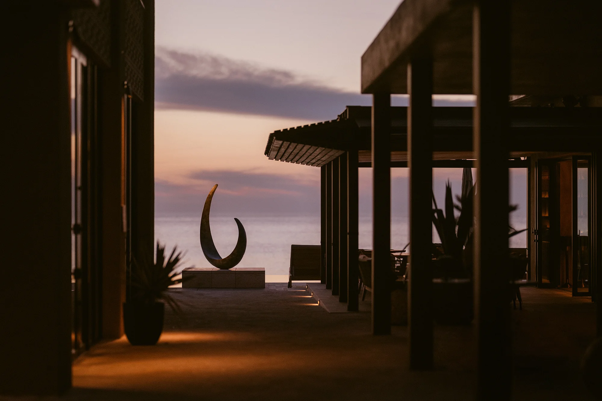 A crescent sculpture glows at the end of a dark colonnade as the sun sets over the sea at Rosewood Miyakojima.