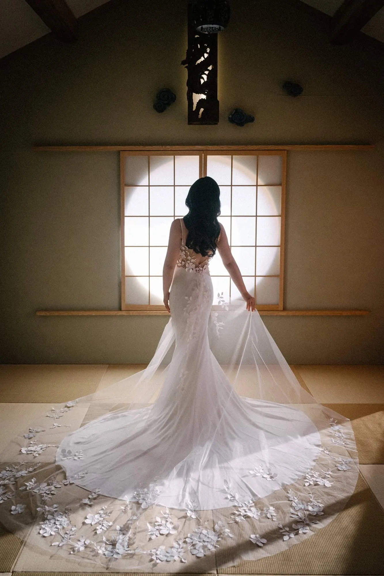 The bride faces a glowing round shoji window, her lace cathedral train fanned across the checkerboard tatami of the villa in Nara.