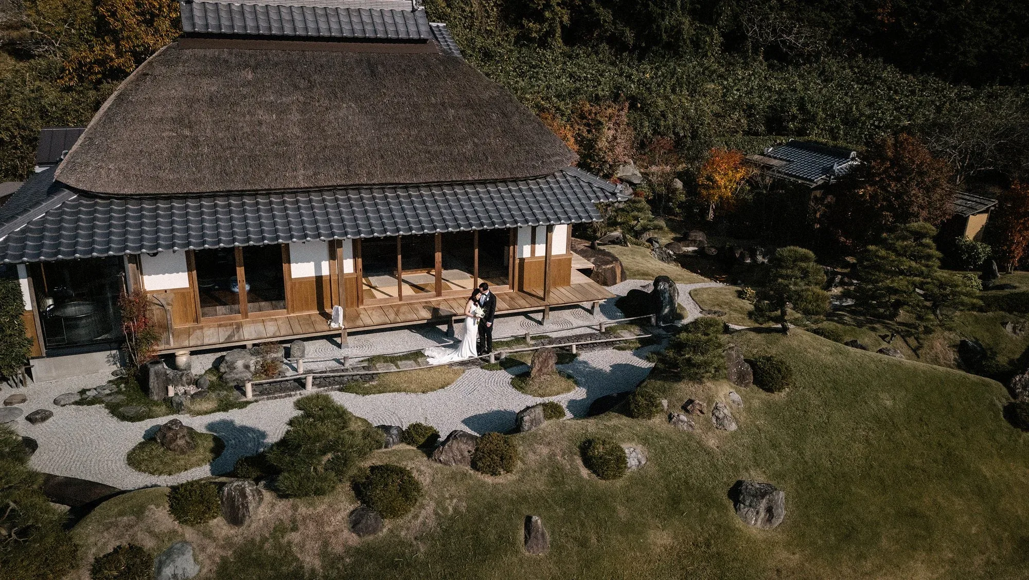 From above, a couple stand on the veranda of a thatched teahouse beside a dry garden.