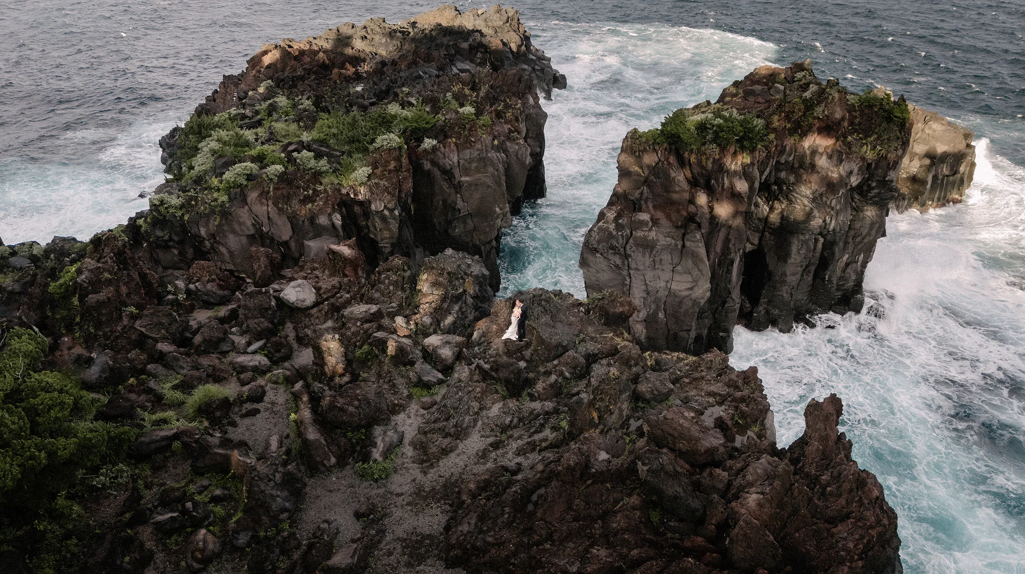 From above, a couple stand small on dark volcanic sea cliffs on the Izu coast as waves break white around them.