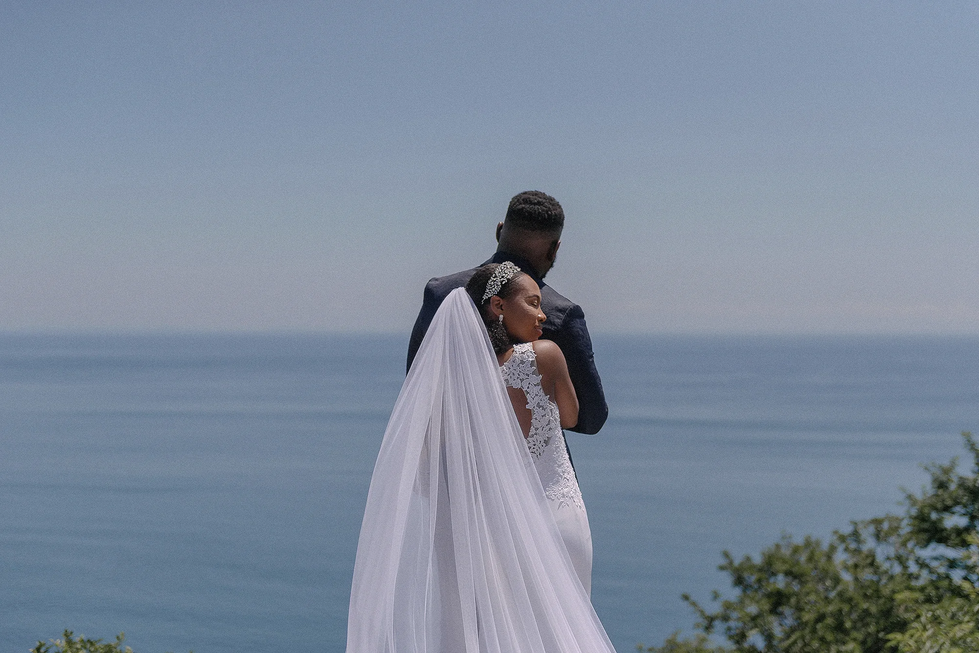 The couple embrace above the calm blue sea at Atami, the bride's long veil falling behind her.