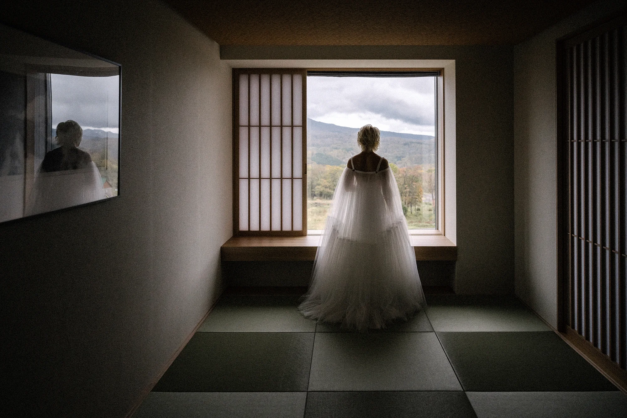 The bride at the end of a dark tatami room, framed by a window onto the hills.