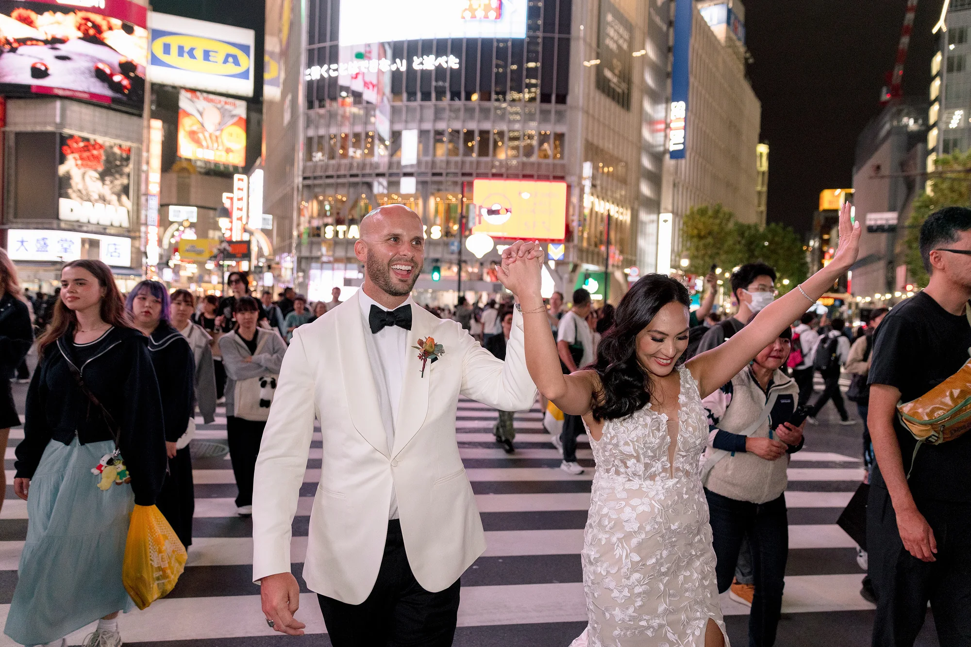 The bride raises her arm in celebration as the couple cross Shibuya at night among the crowd.