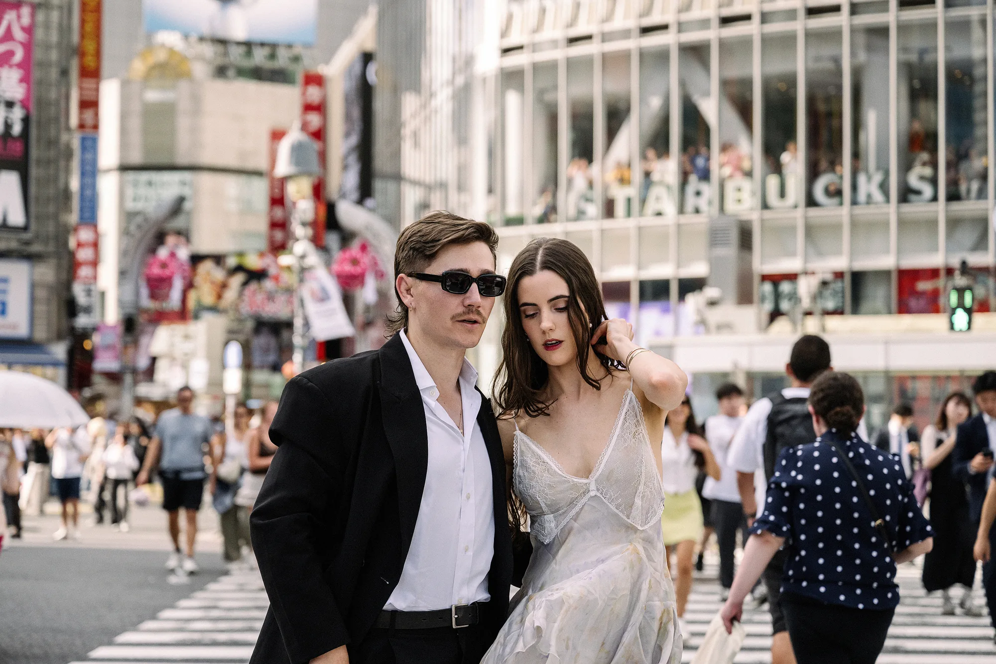 The groom in sunglasses and a black jacket walks arm in arm with the bride in a pale slip dress across the Shibuya scramble crossing.