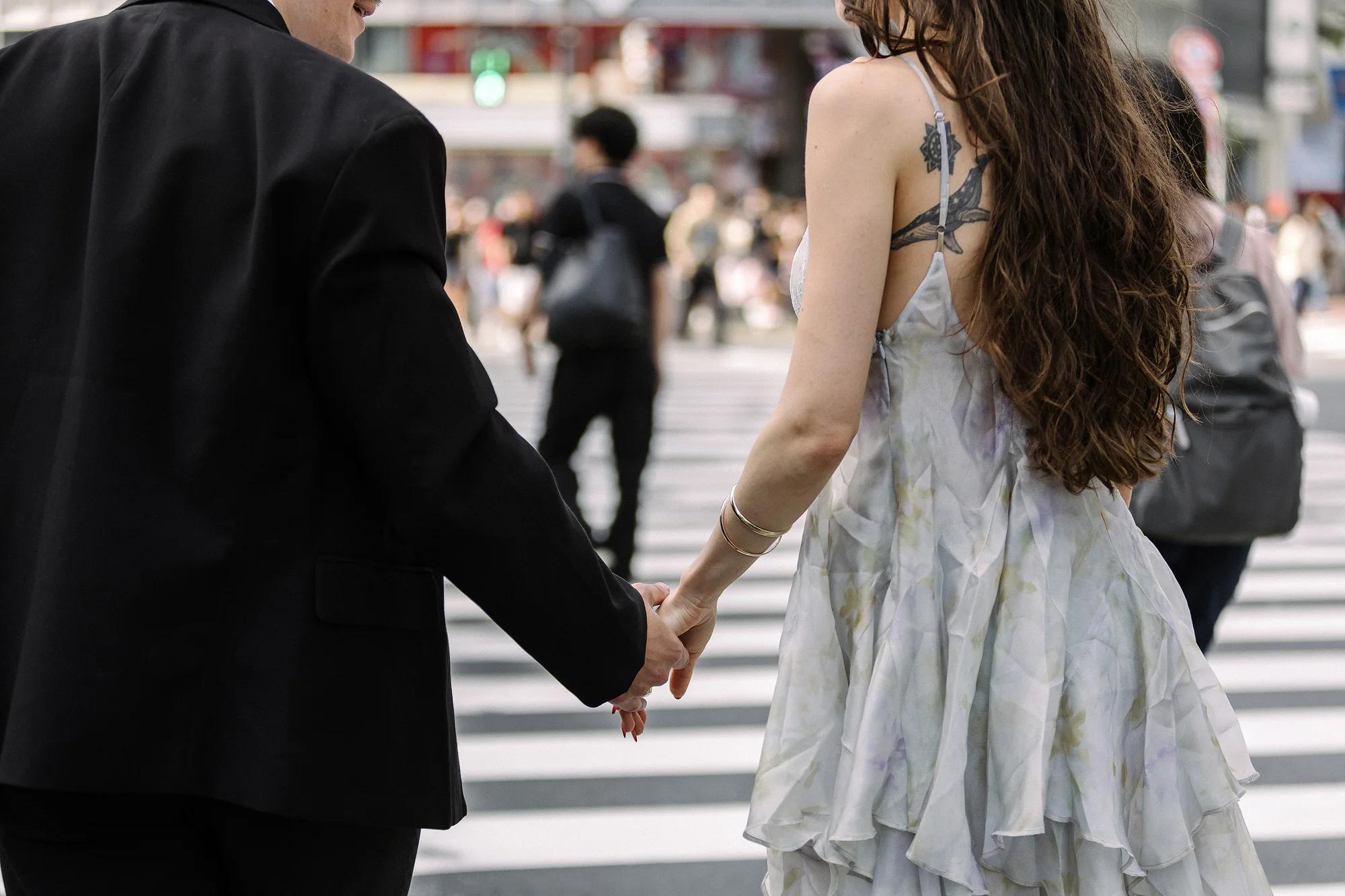 A couple walk hand in hand across the Shibuya crossing, crowds all around them.