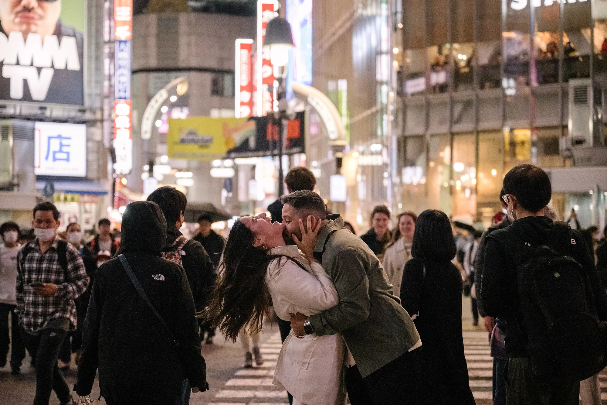 A couple kiss in the middle of the Shibuya crowd at night, neon signs above.