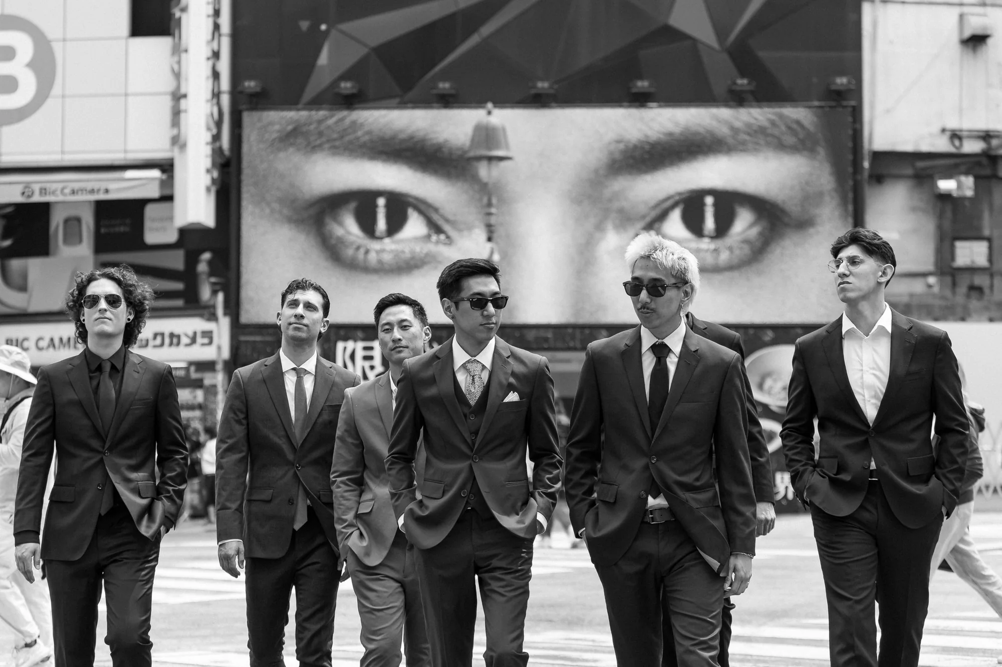 In black and white, the groom and his groomsmen walk in sunglasses beneath a giant billboard of eyes in Shibuya.