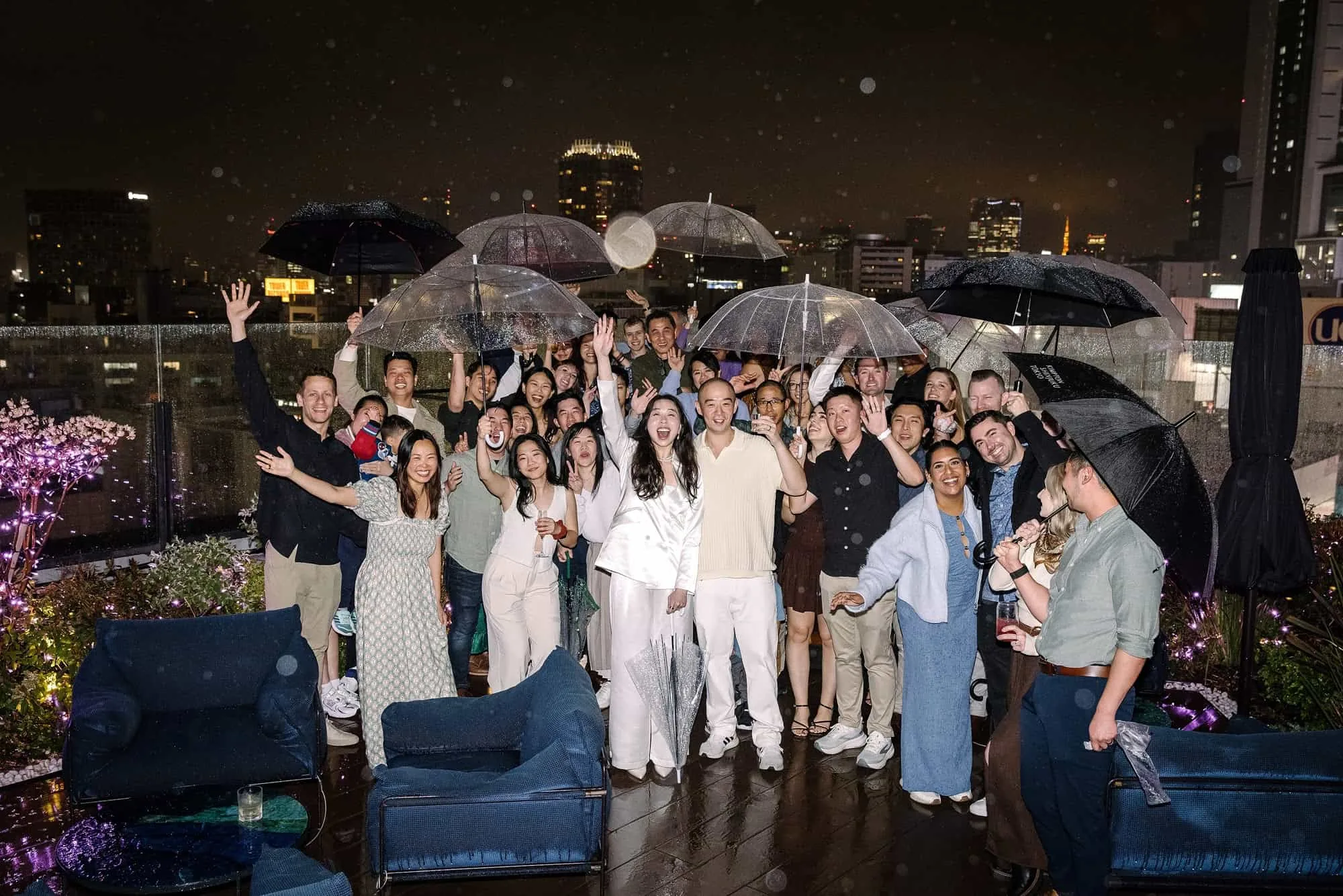 Guests crowd together under umbrellas on a rainy Shibuya rooftop at night, cheering at the start of a destination wedding weekend in Japan.