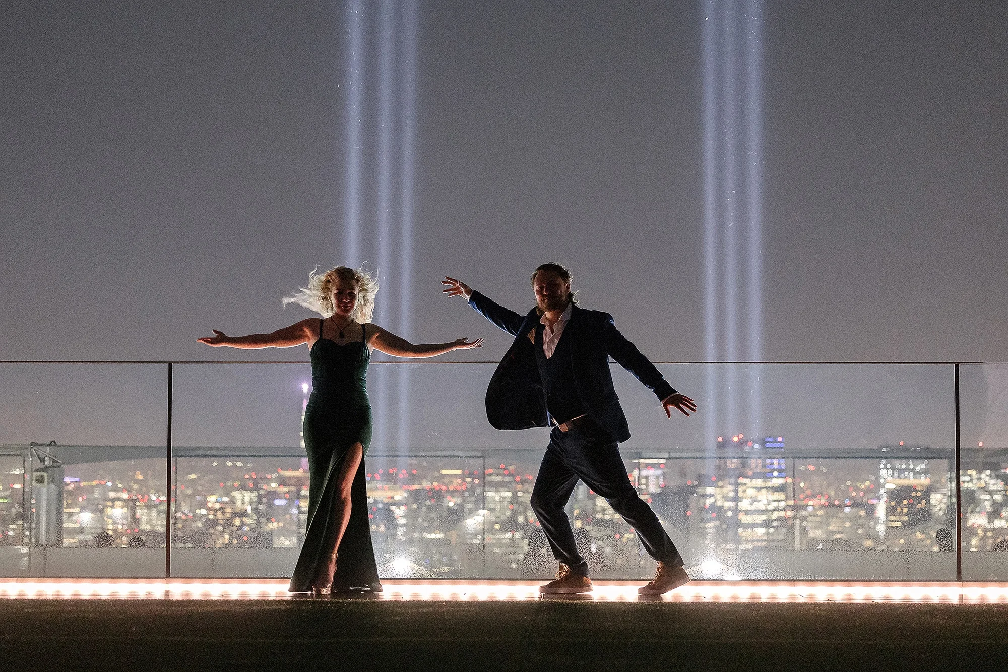 A couple dance with arms outstretched on a Tokyo rooftop at night, beams of light rising behind them.