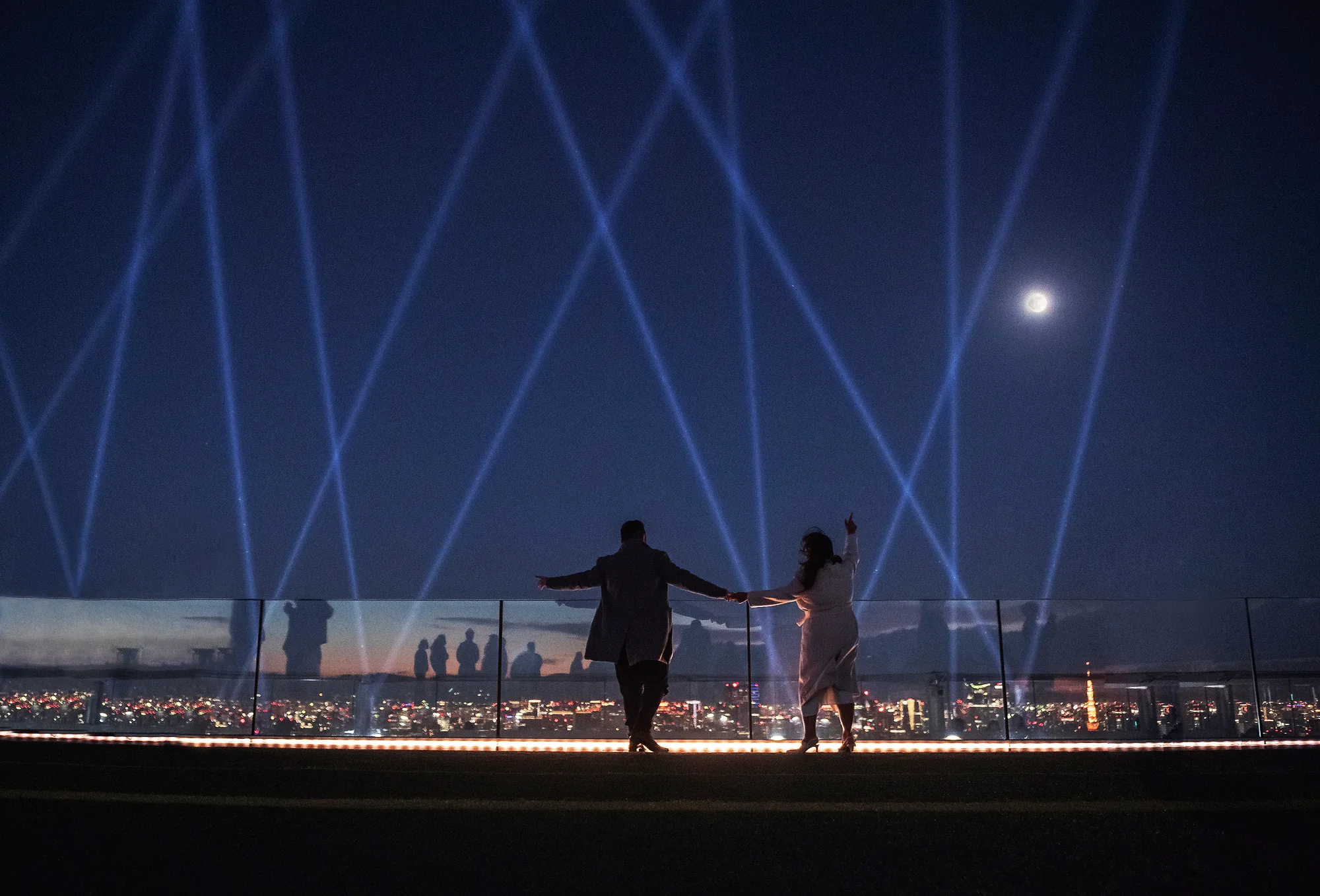 The couple reach for each other on a rooftop at night as beams of light cross the sky above Shibuya.