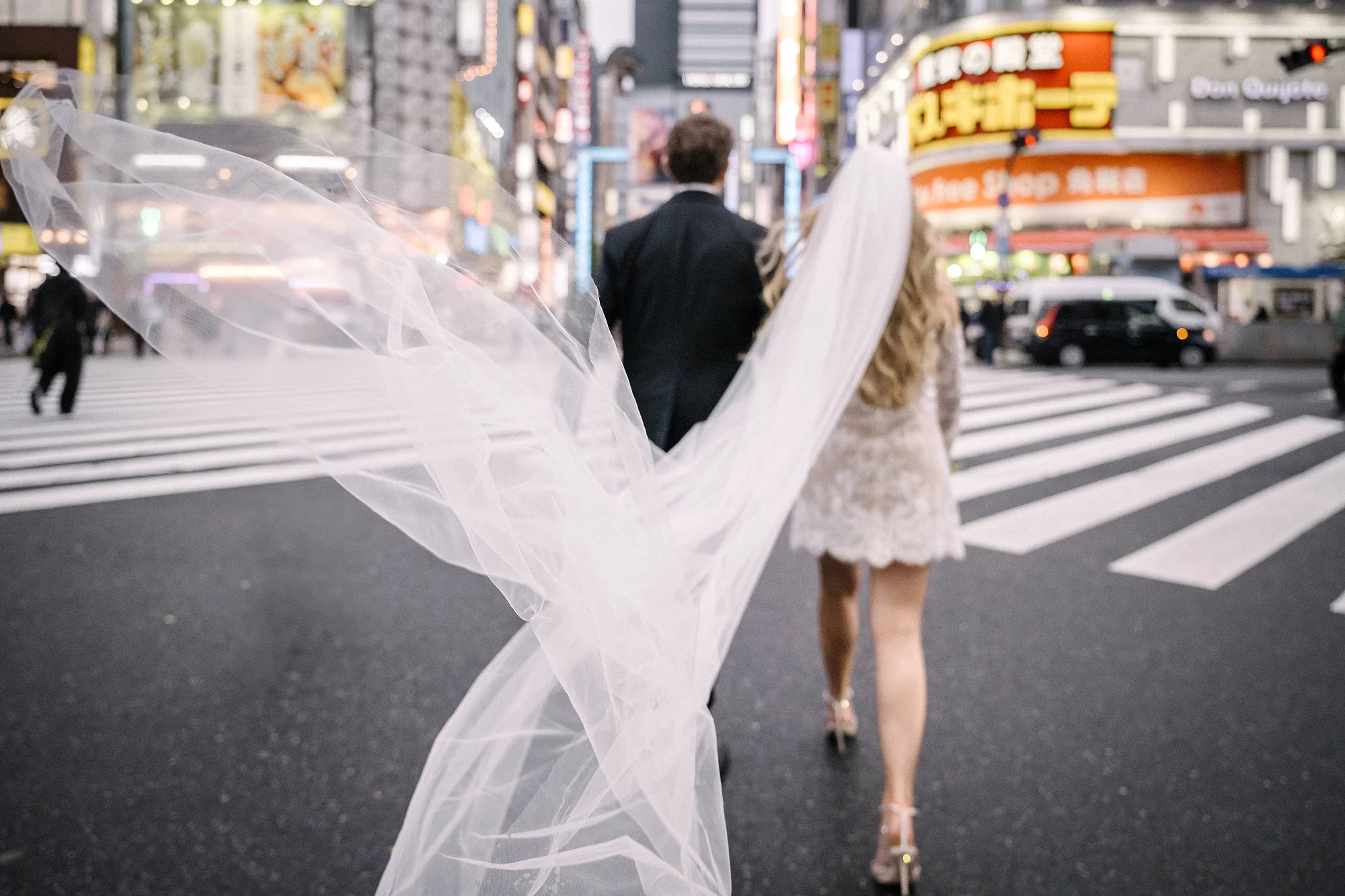 A bride's long veil streams behind her as the couple cross a Shinjuku street, neon signs above.