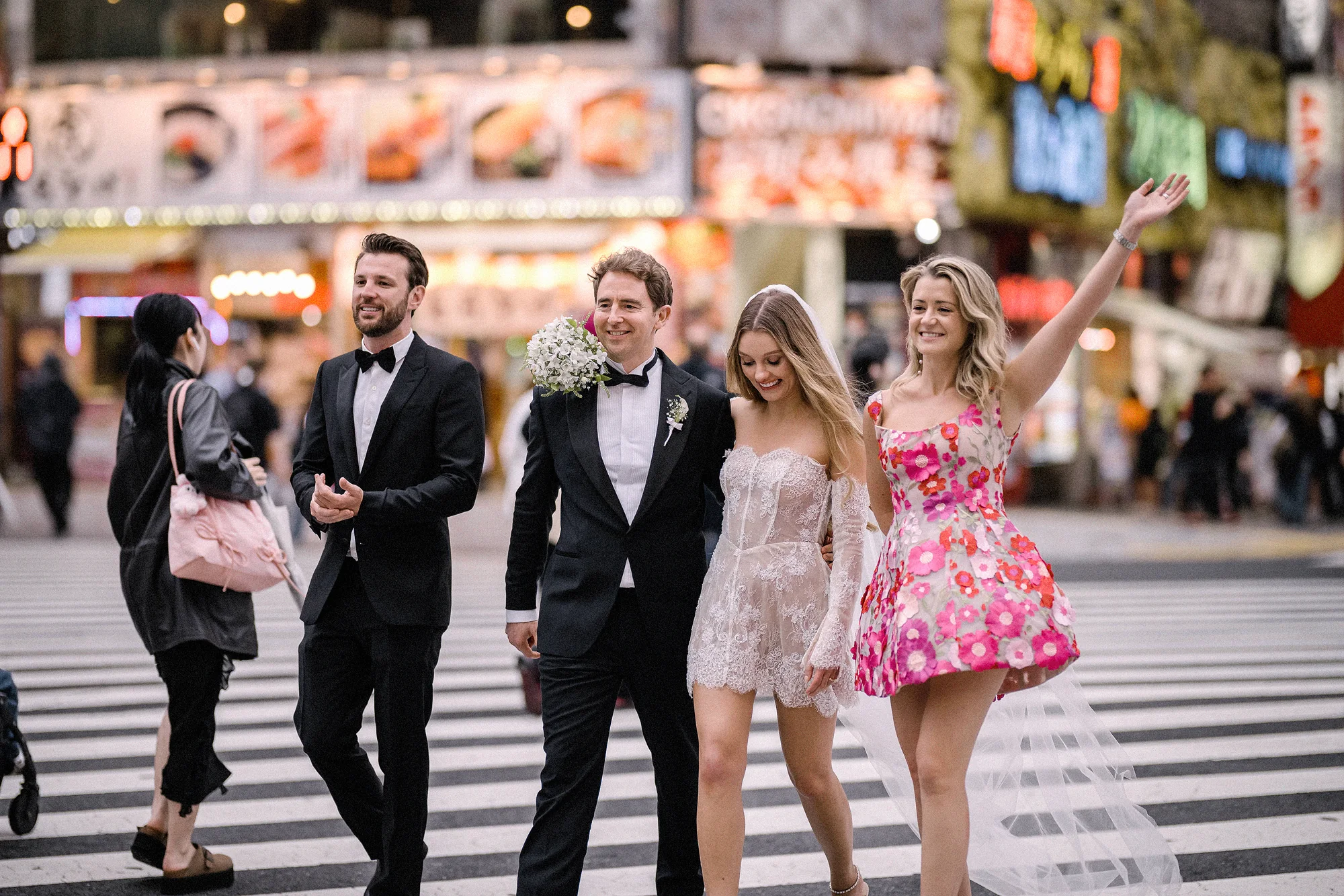 The couple cross a Shinjuku street with their friends, a bridesmaid in a pink floral dress waving, neon signs behind them.