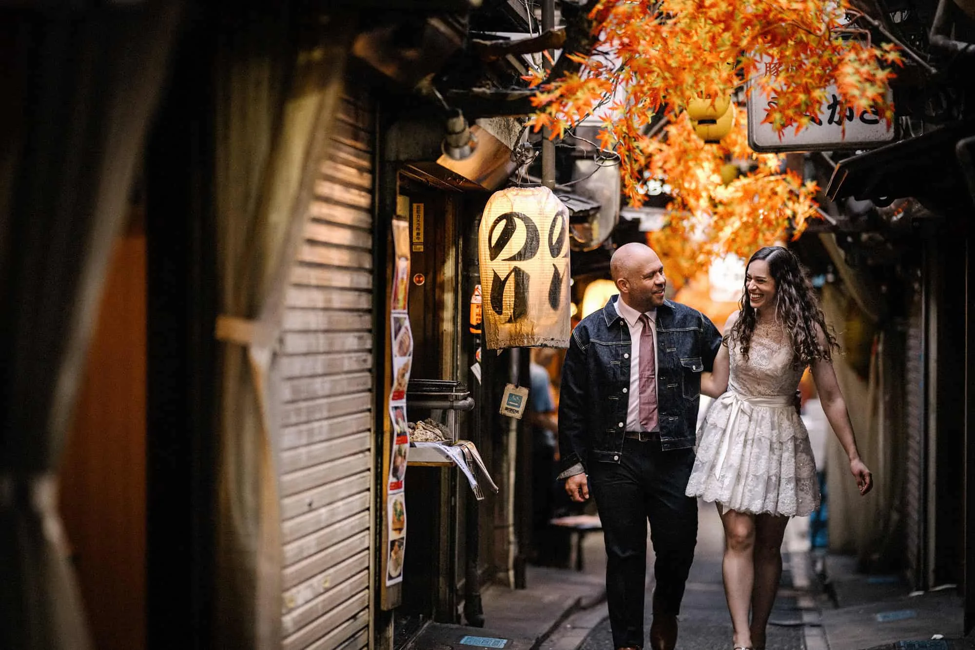 The couple walk laughing down a lantern alley in Shinjuku under paper autumn maple leaves, she in a short lace dress and he in a denim jacket.