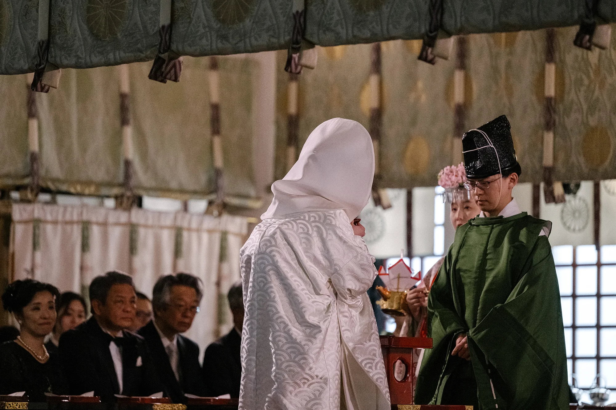 During a Shinto ceremony the bride in a white wataboshi sips sake beside a priest in green robes, family seated behind.