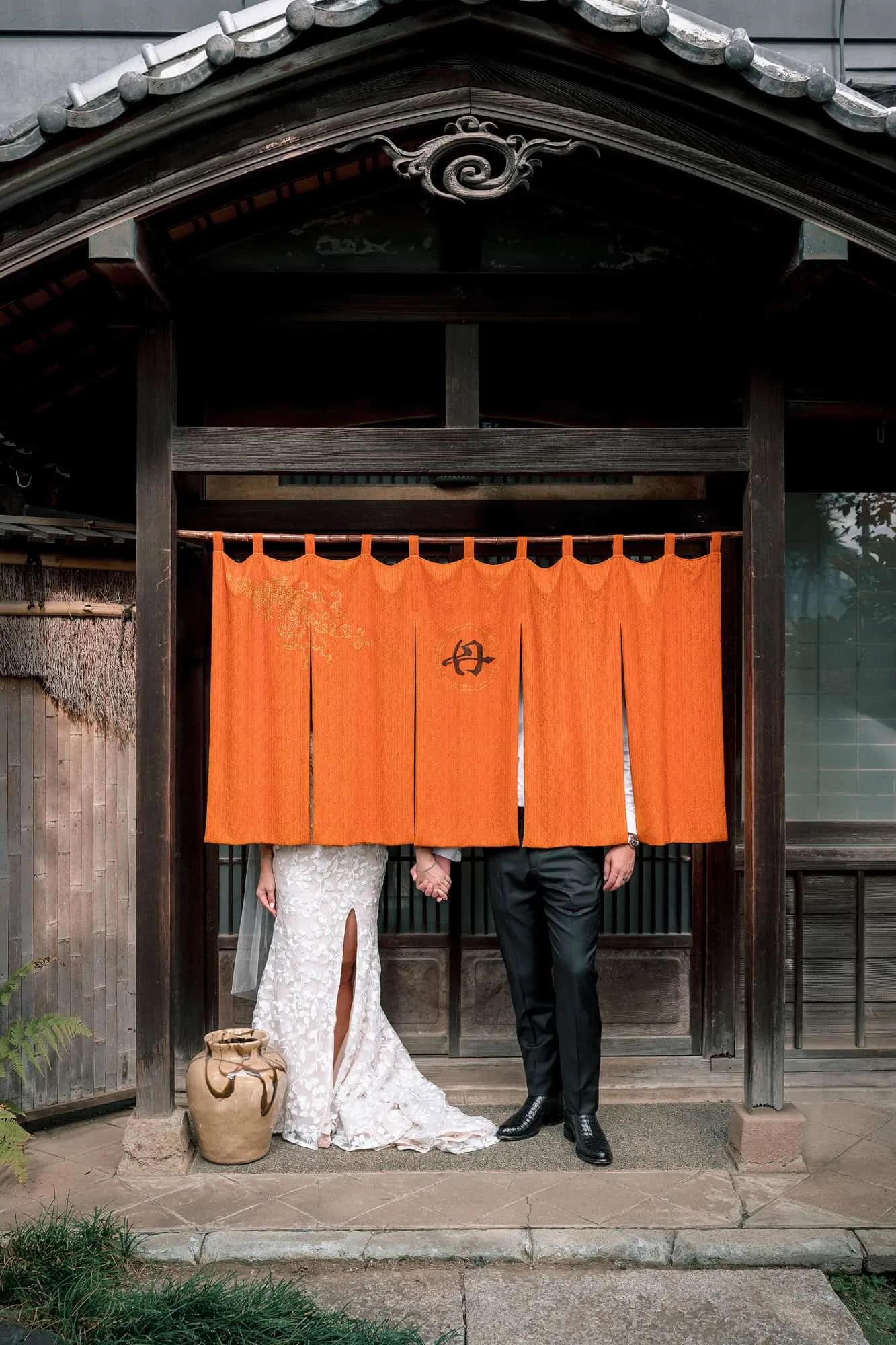 The bride and groom peek out from under an orange noren marked with the house crest at the timber doorway of the old house at Tantoku.