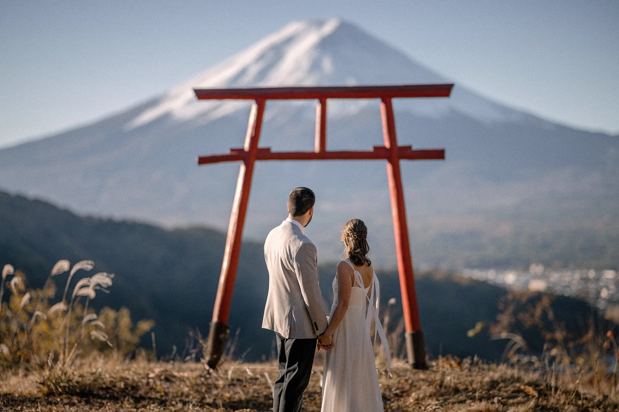 A couple stand beneath a red torii gate high on a hillside, Mt Fuji beyond.