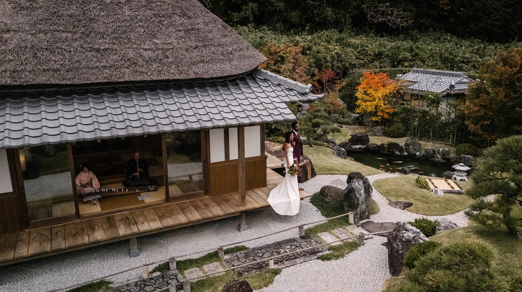 The couple on the veranda of a thatched teahouse above a raked dry garden in autumn.