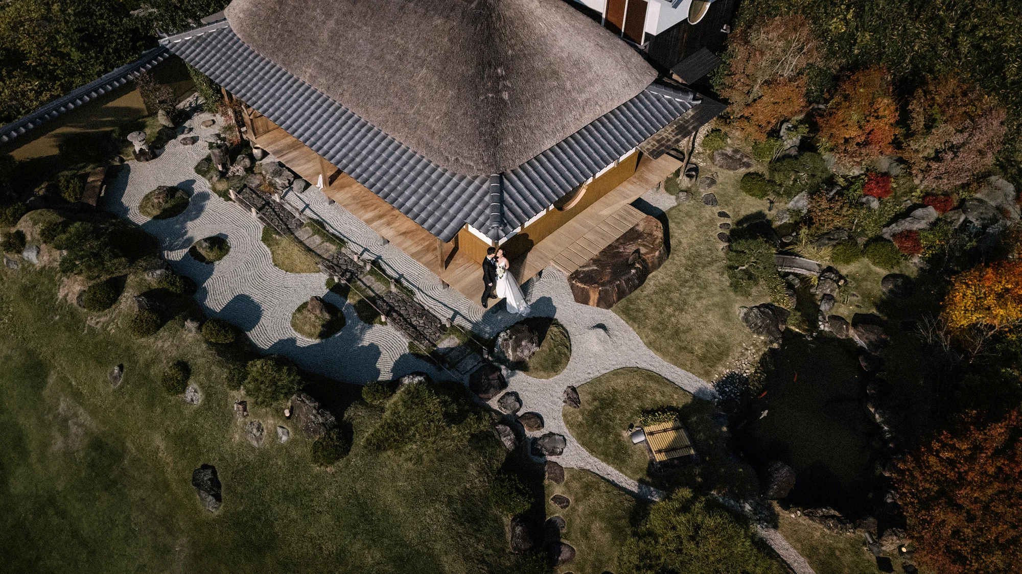 From above, the thatched roof of a villa and its stone garden, the couple small on the path.