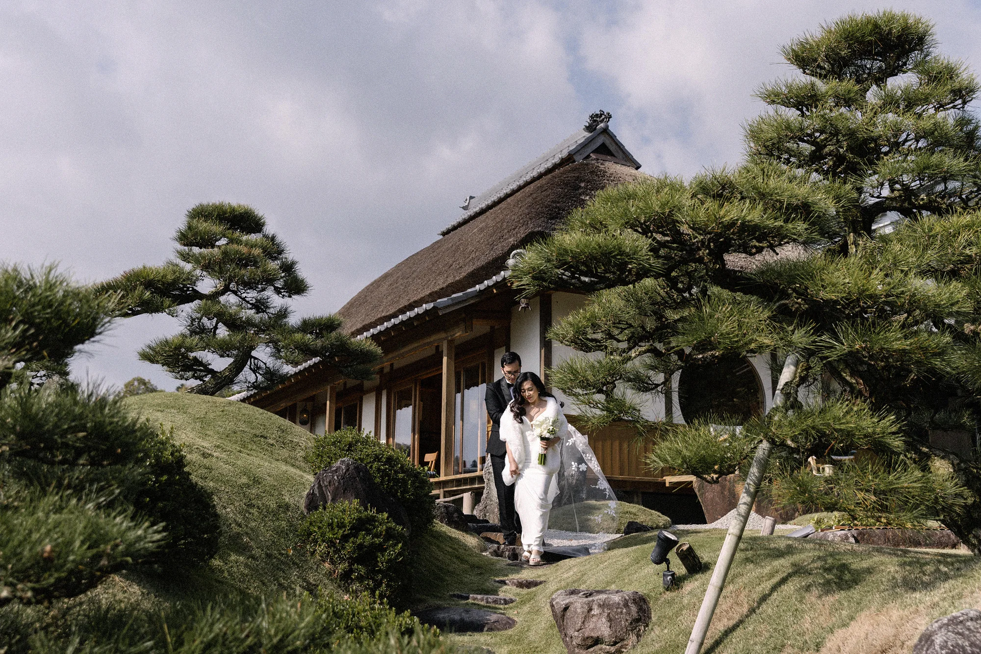 A couple walk through a pine garden below a thatched villa.