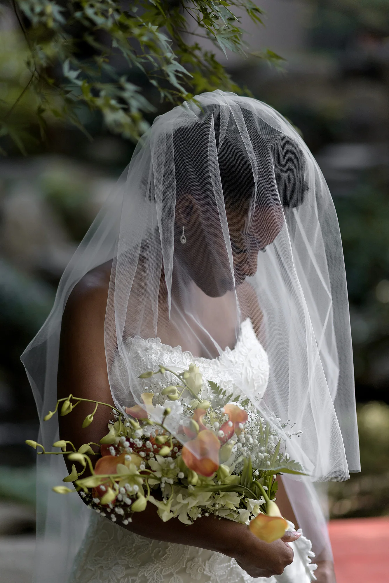 The bride looks down through her veil at a bouquet of orange and white flowers in Tokyo.