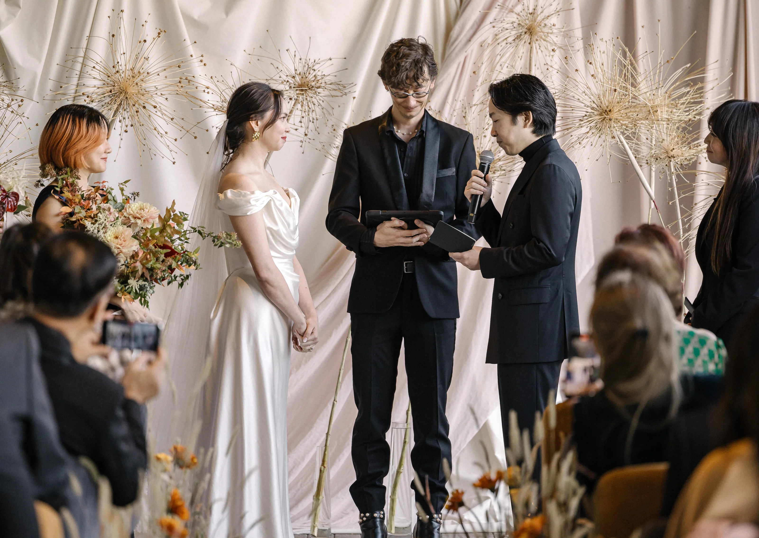 A couple stand before an installation of dried flowers and pale drapes as their friend leads the ceremony in Tokyo, guests looking on.