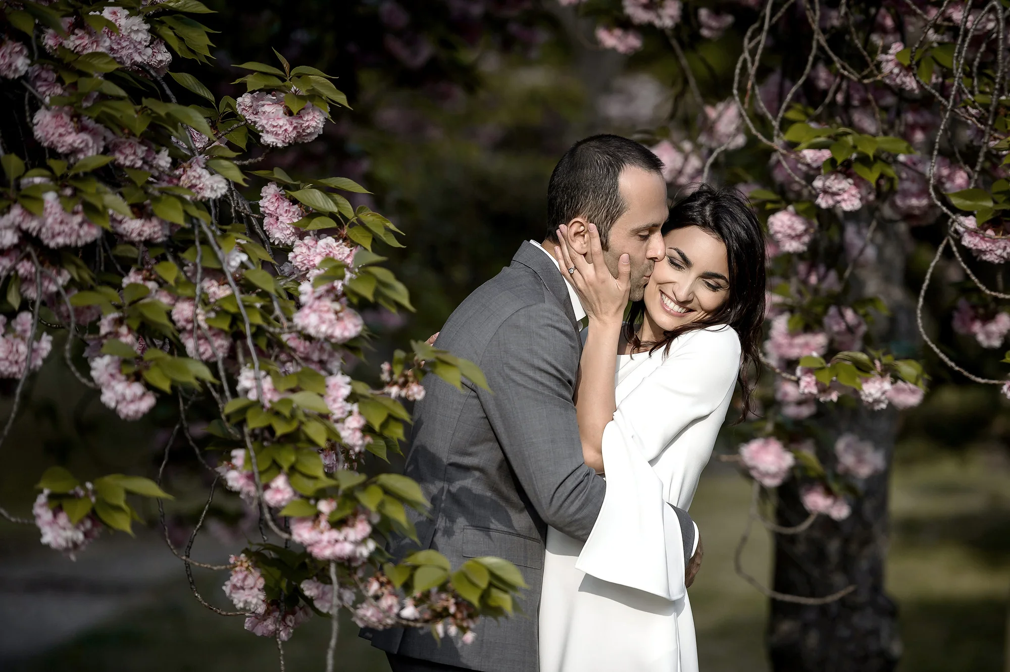 The groom kisses the bride's cheek beneath pink cherry blossom in Tokyo.