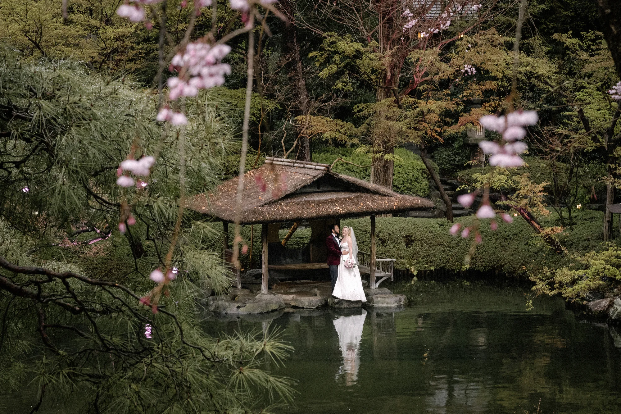A bride and groom by the pond pavilion of a Tokyo garden in spring, blossom in the foreground.
