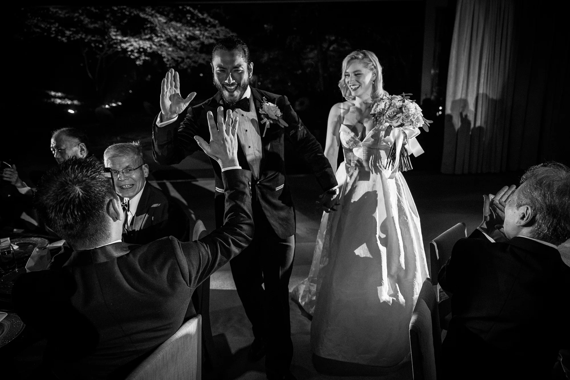 In black and white, the groom high-fives a guest as the couple make their entrance at their Tokyo reception, the bride laughing beside him.