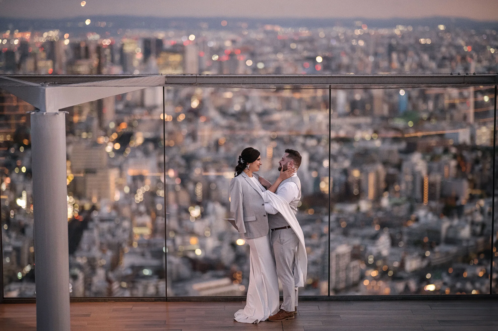 A couple embrace behind glass on a Tokyo rooftop at sunset, the city spread below.