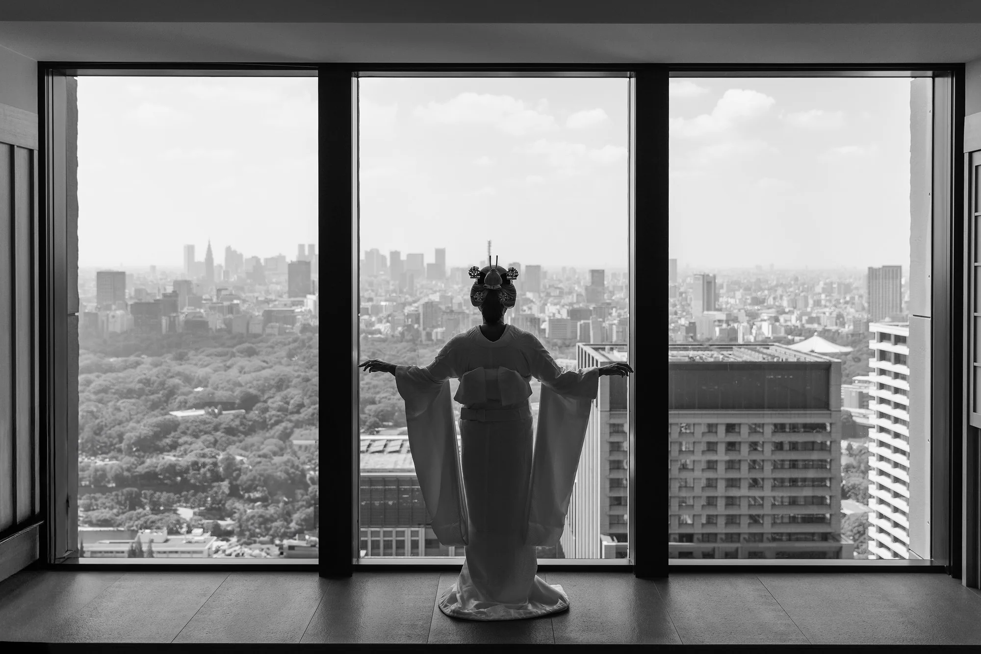 In black and white, a bride in kimono stands at a tall window high above the Tokyo skyline.