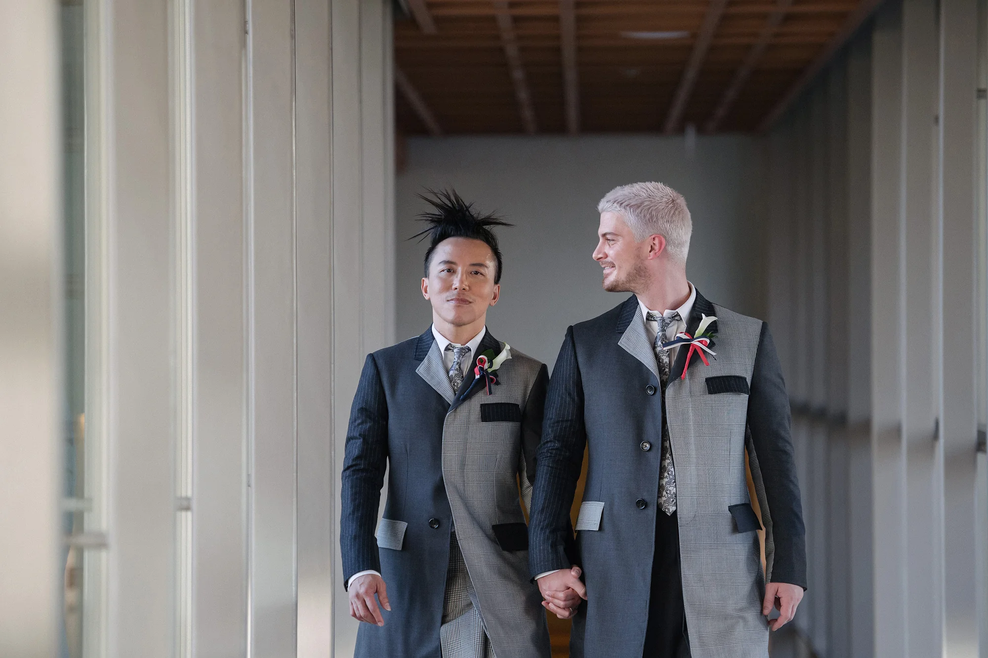 Two grooms in gray suits hold hands and smile at each other in a pale colonnade in Tokyo.