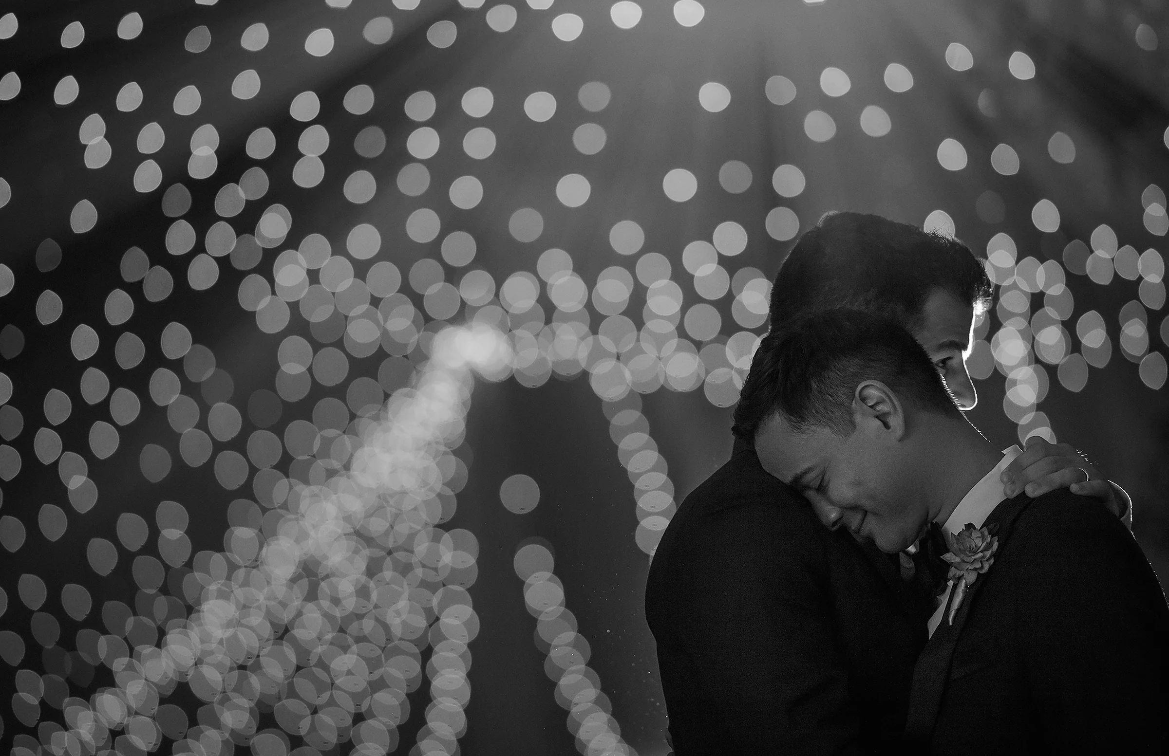 In black and white, two grooms hold each other close under a canopy of soft lights.