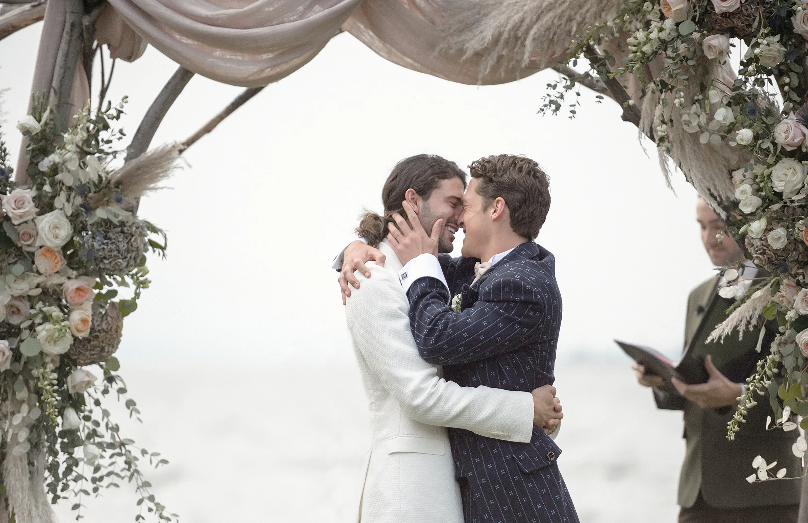 Two grooms embrace beneath a floral arch at their ceremony as the celebrant looks on.