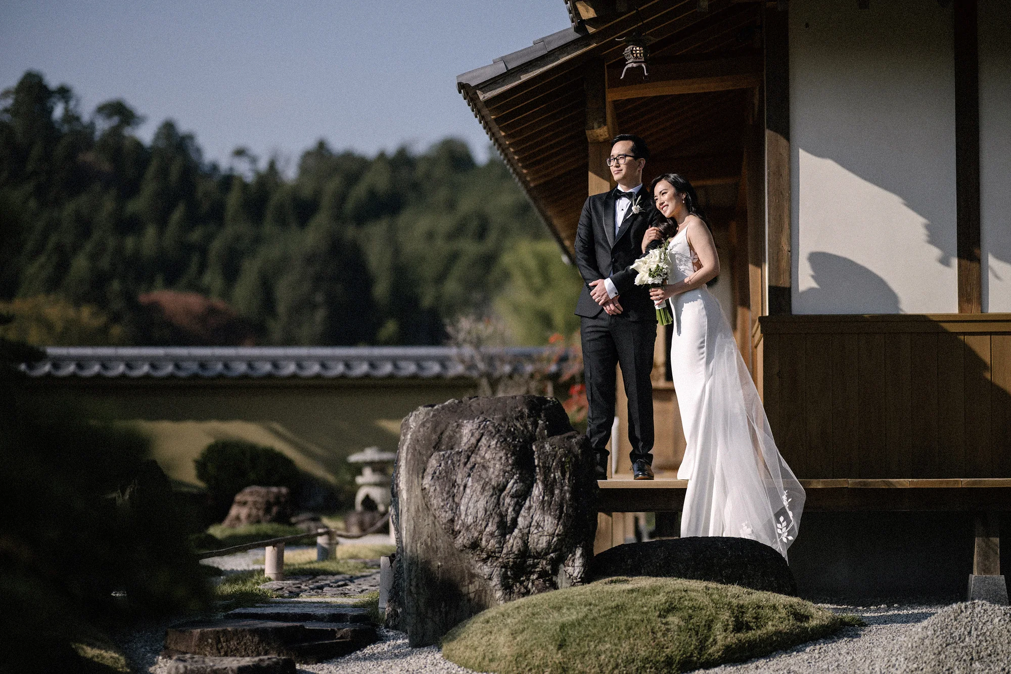 The couple stand on the step of a villa veranda above a Japanese garden.