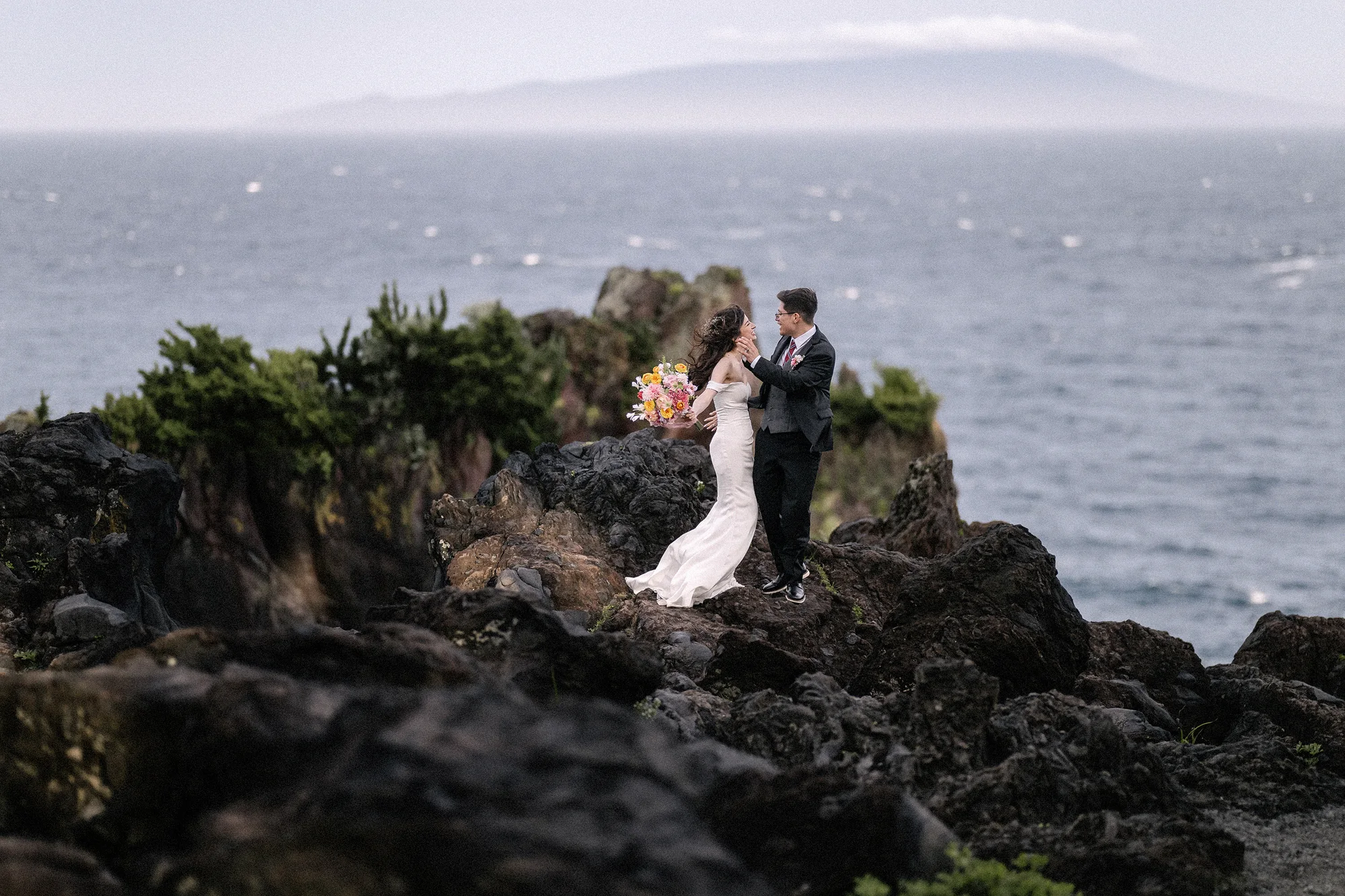 A couple stand on dark volcanic rocks above the sea on the Izu coast, an island on the horizon.