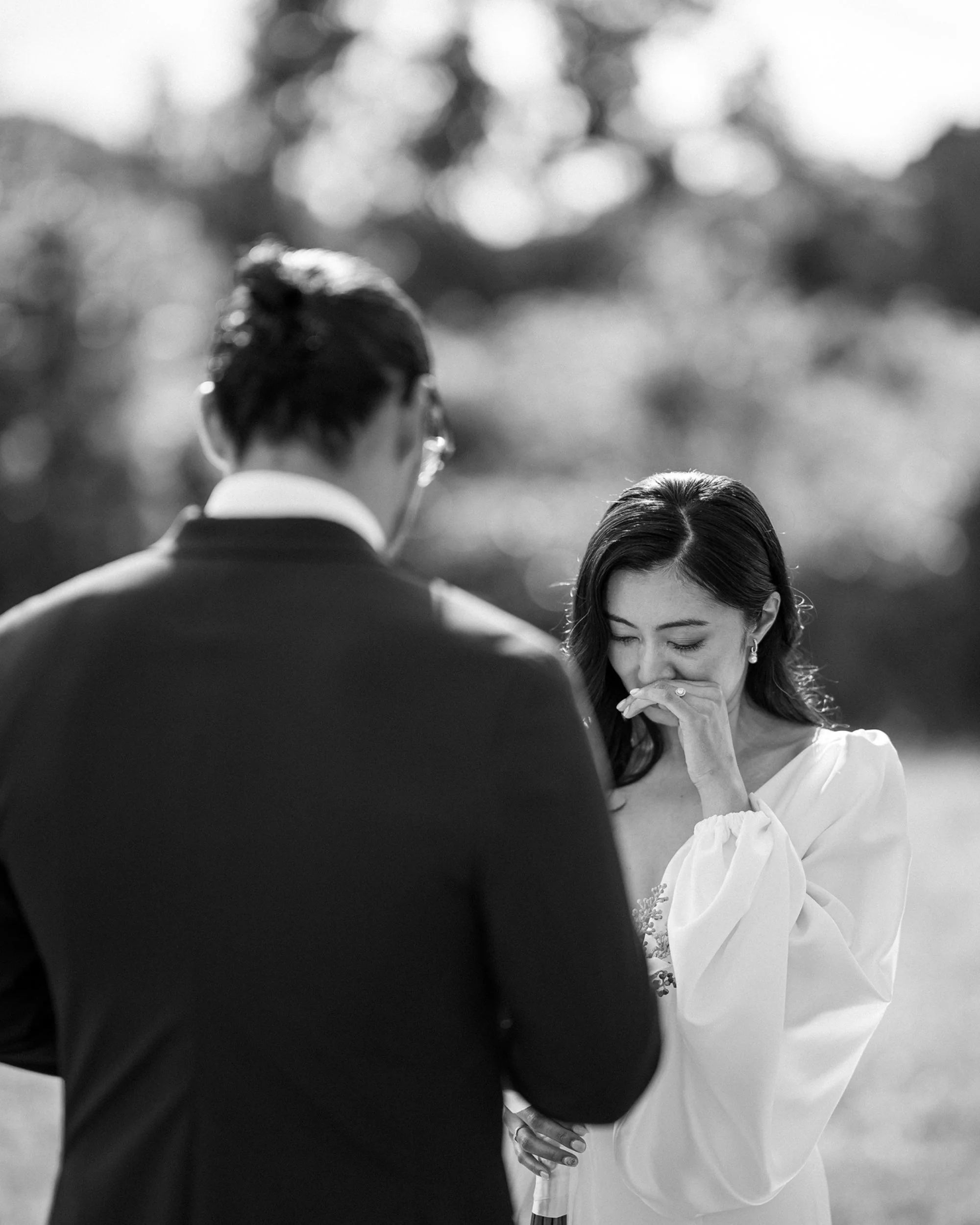 In black and white, the bride covers her mouth, moved to tears, as the groom reads his vows.