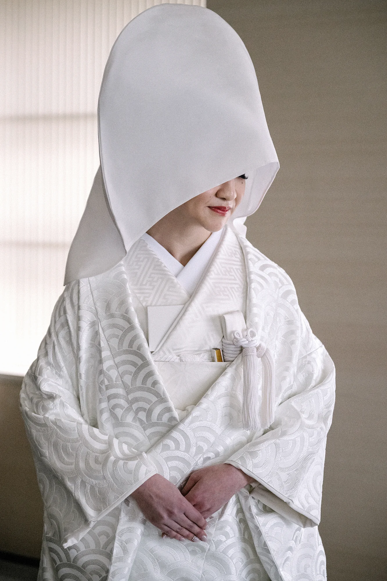 The bride sits in a white wataboshi hood and shiromuku, hands folded, in soft window light.