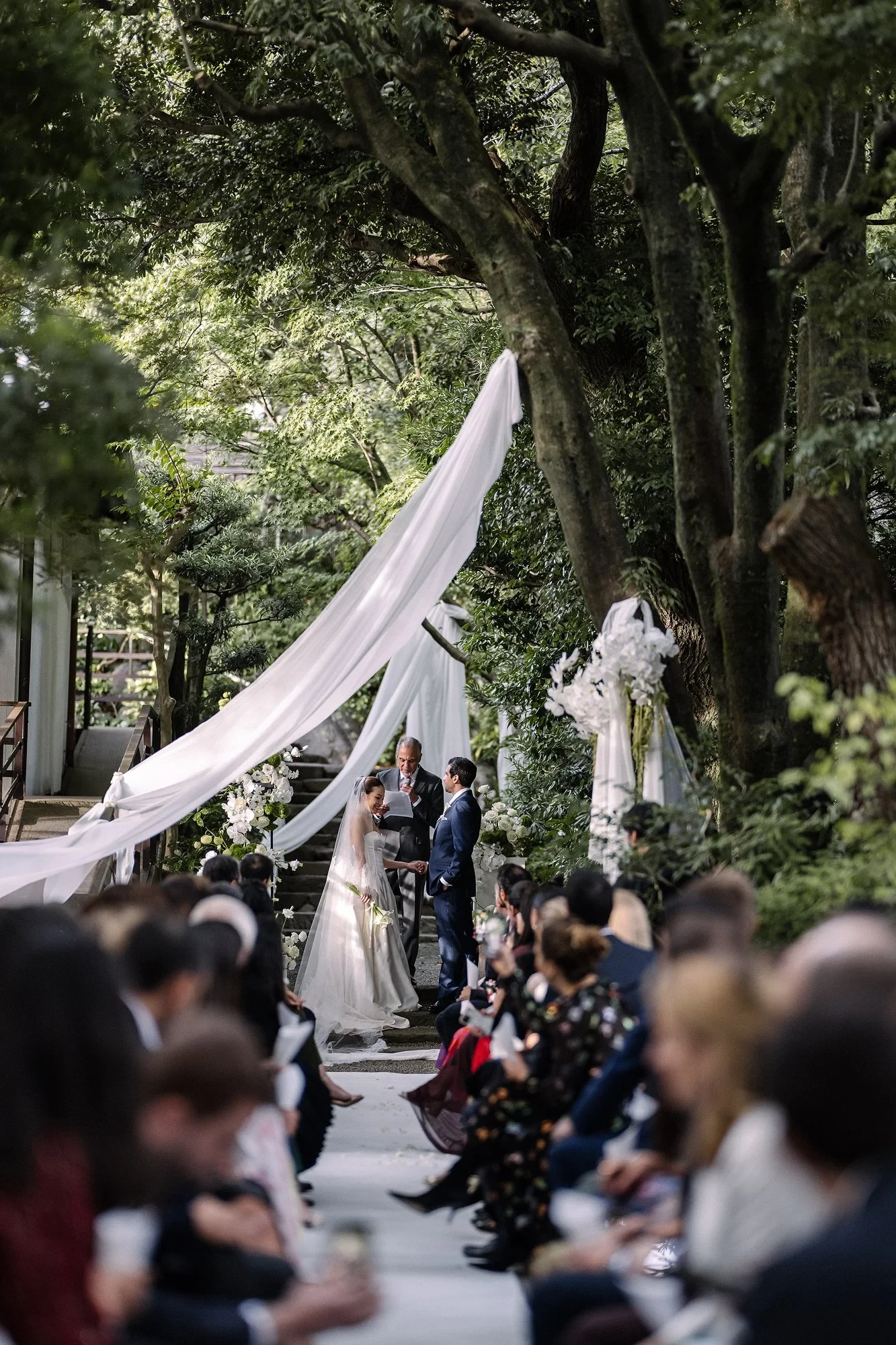 A couple hold hands at an outdoor ceremony, with guests seated beneath trees.