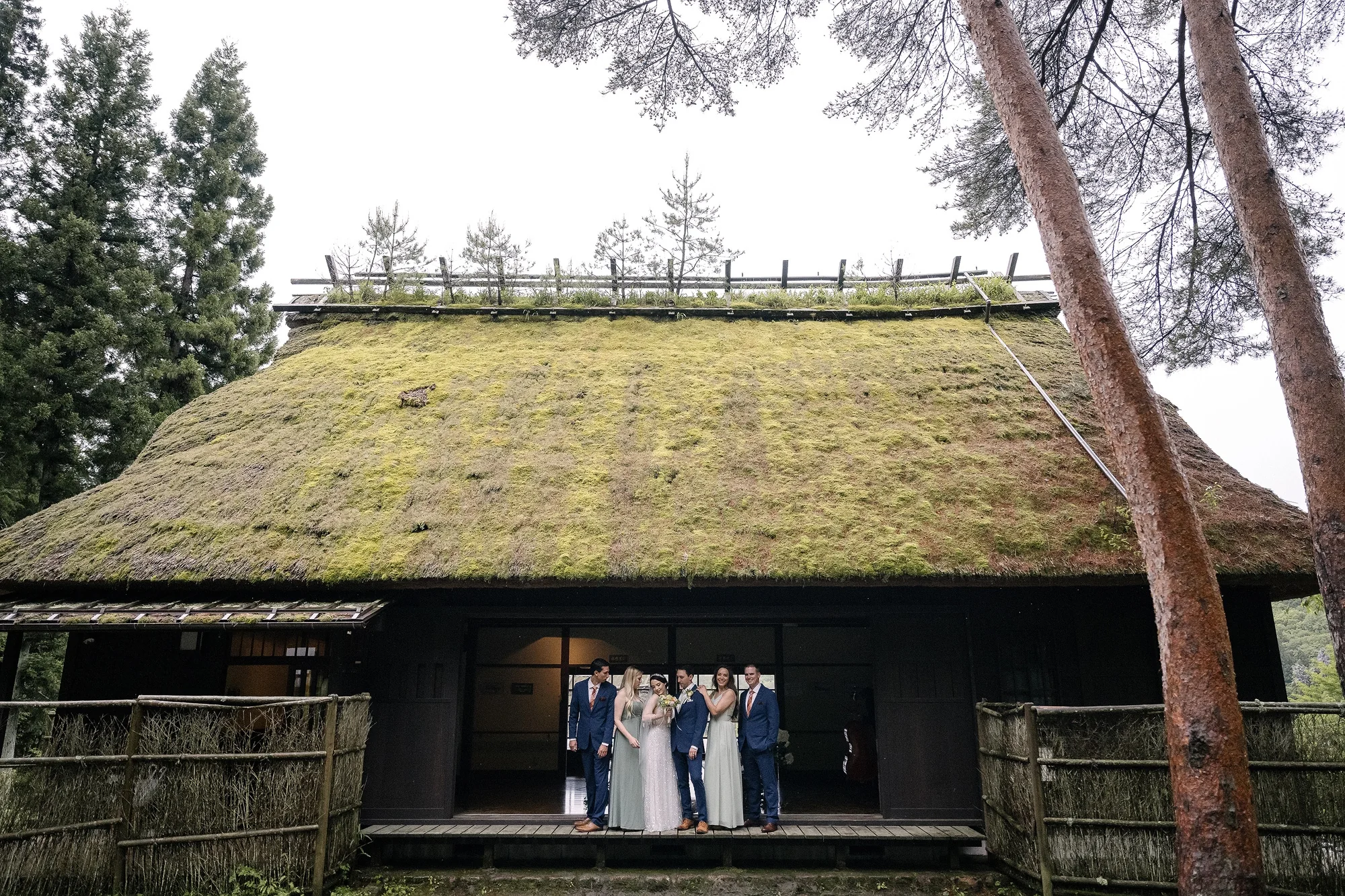 The wedding party gather outside a thatched farmhouse roofed in moss, tall cedars around them.