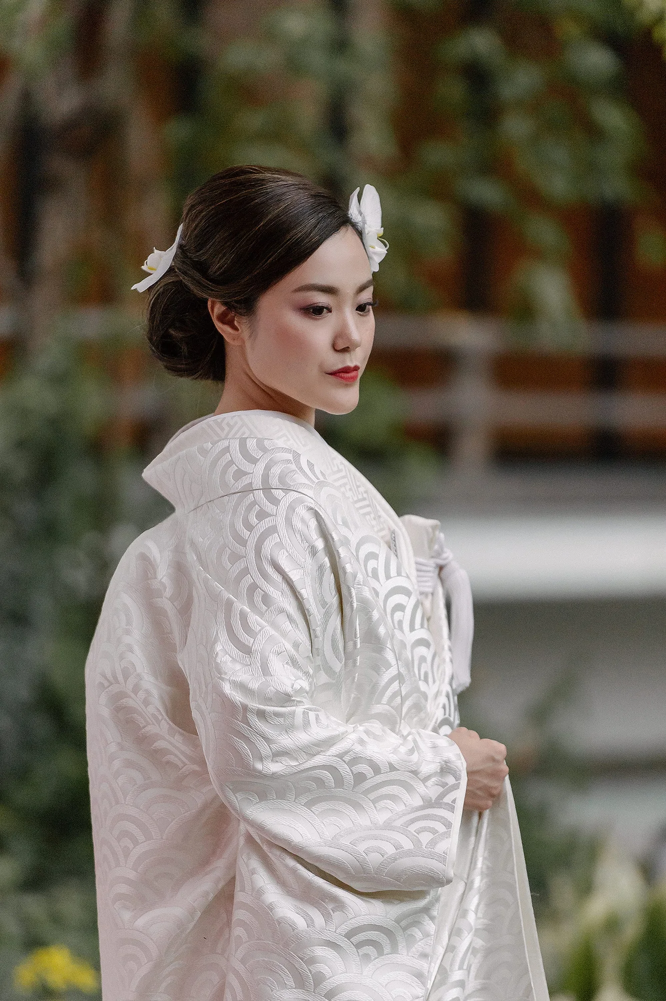 The bride in a white kimono woven with a wave pattern, orchids in her hair, in a garden.