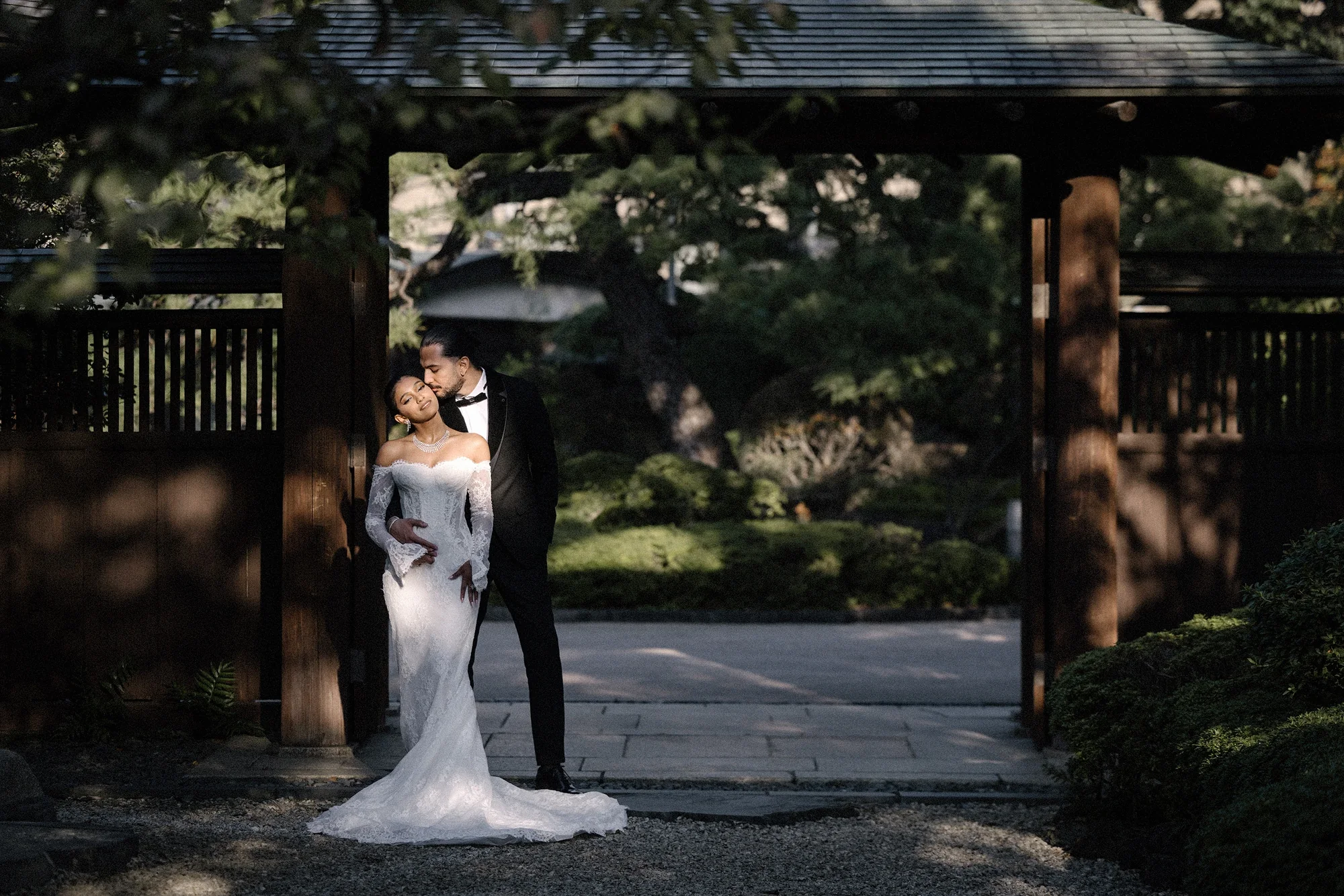 The couple kiss beneath a wooden gate opening onto a Japanese garden.
